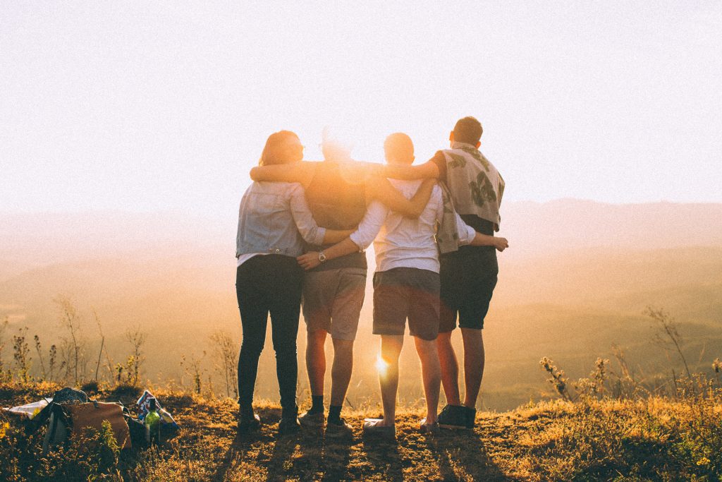 Four friends standing with their arms around each other, looking out at a sunrise over the hills