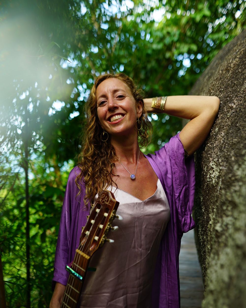 Francie La Flow smiling with her guitar against a jungle rock wall